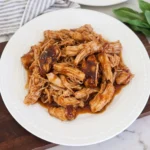 Tender shredded Crockpot BBQ Chicken coated in glossy smoky barbecue sauce on a white serving plate